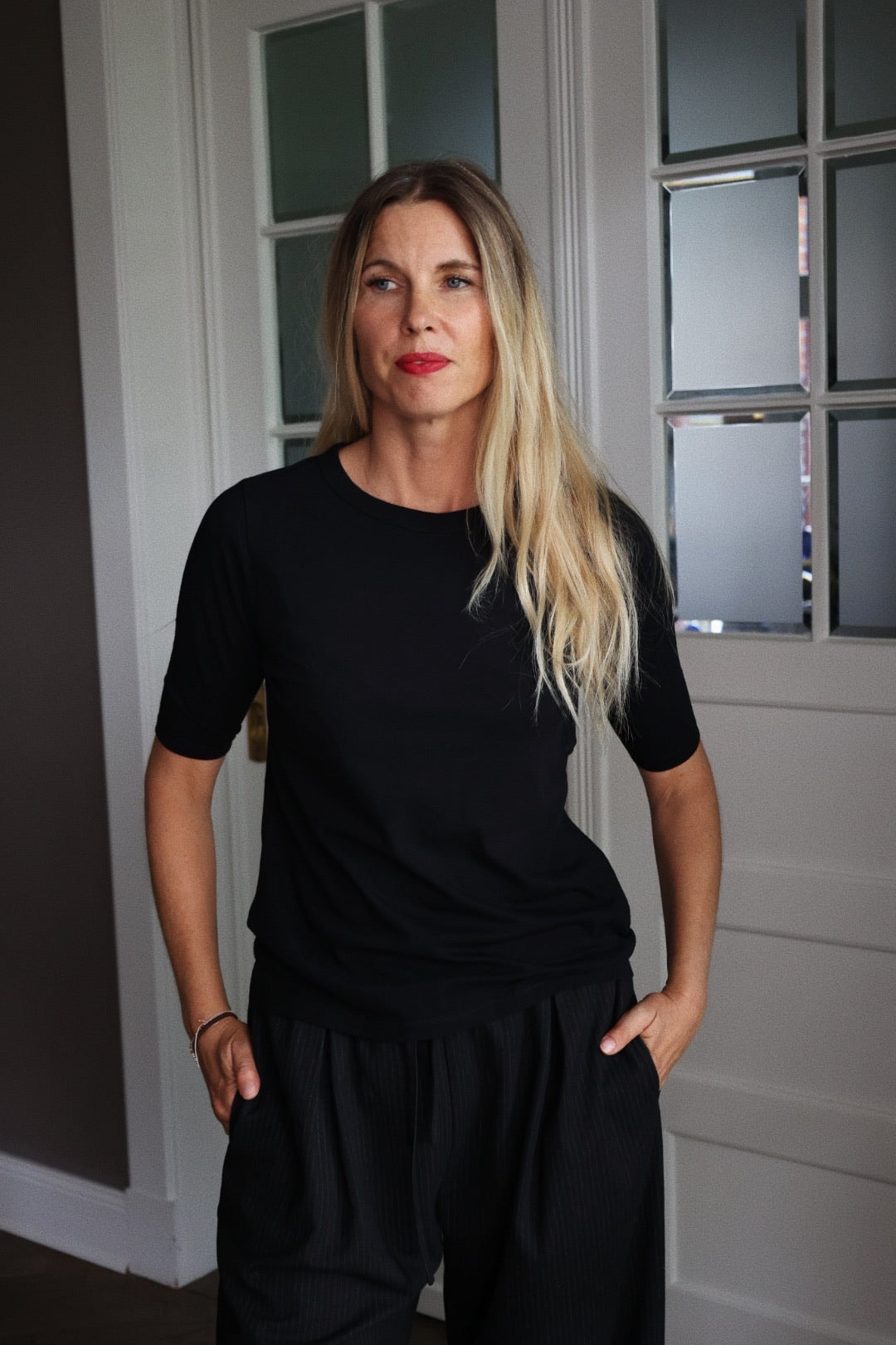 Eine Frau mit langen blonden Haaren und rotem Lippenstift steht in einem Innenraum, trägt ein schwarzes T-Shirt mit halben Ärmeln und eine schwarze Hose, hat die Hände in den Taschen und posiert vor einer weißen Tür mit Milchglasscheiben.