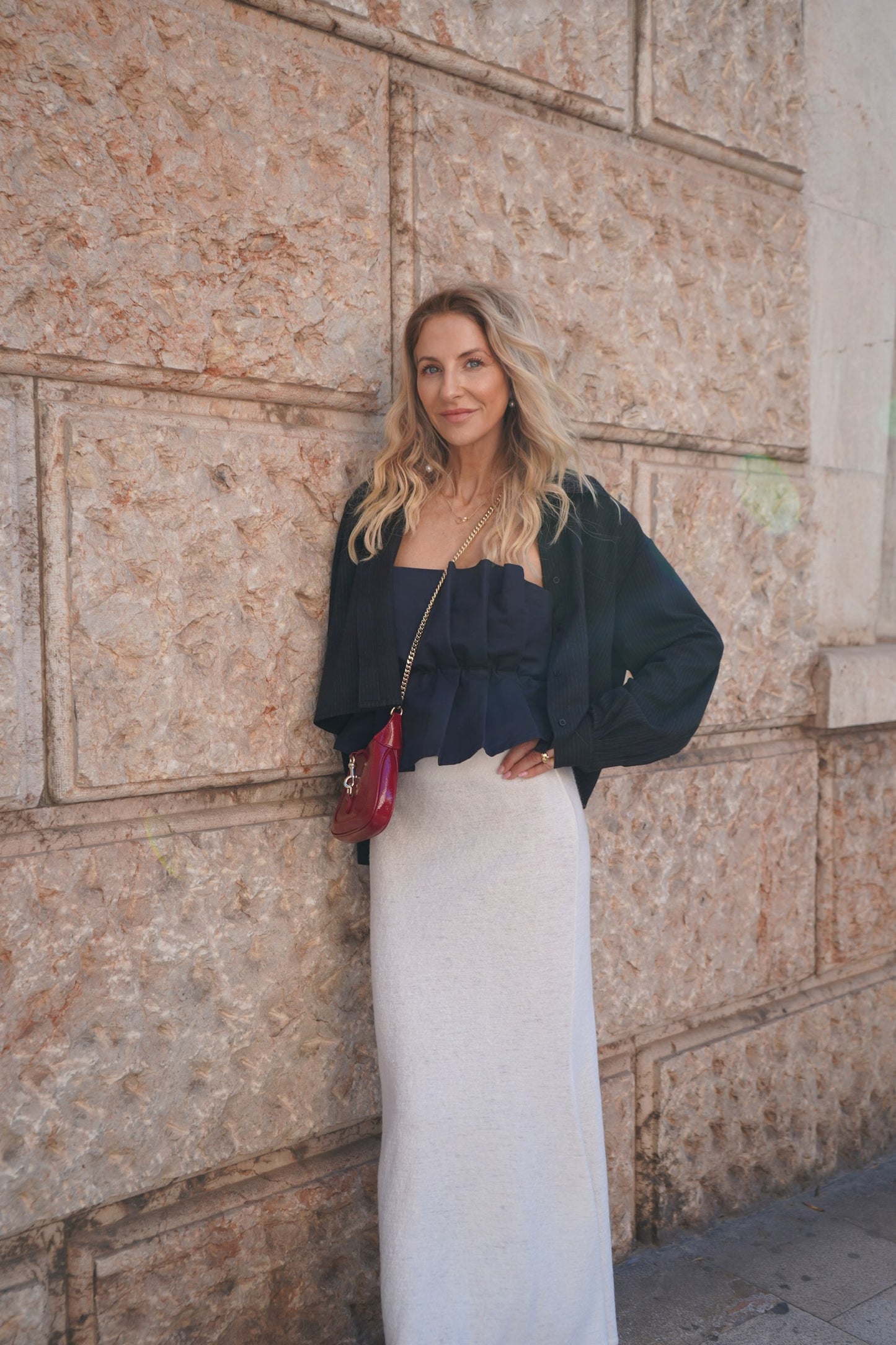 Eine Frau mit gewelltem blondem Haar steht vor einer strukturierten Steinmauer. Sie trägt das Pleat Top Navy, einen langen cremefarbenen Rock, eine schwarze Jacke und eine kleine rote Handtasche, die sie über die Schulter hängt. Sie blickt mit einem leichten Lächeln in die Kamera.