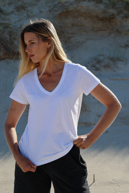 Eine Frau mit langen blonden Haaren steht im Freien auf sandigem Boden, trägt ein weißes T-Shirt mit V-Ausschnitt und eine schwarze Hose und schaut mit einer Hand in der Hüfte zur Seite.