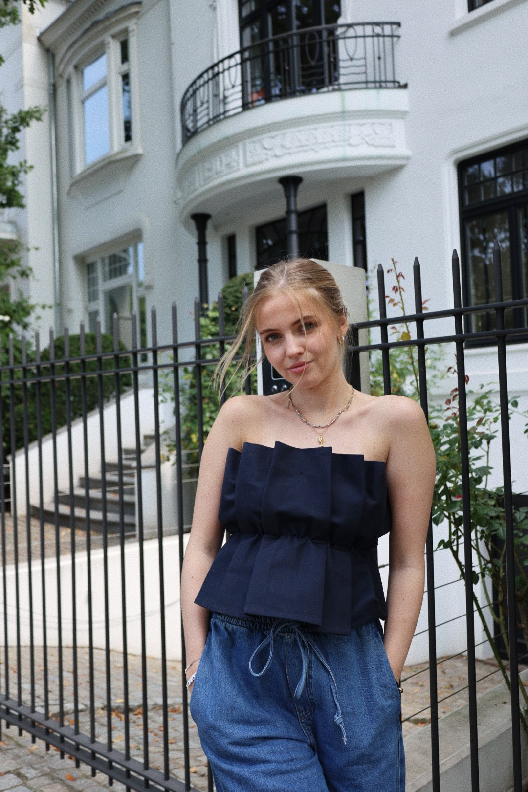 Eine junge Frau mit blondem Haar steht vor einem schwarzen Eisenzaun. Sie trägt ein modisches Pleat Top Navy und eine blaue Jeans, im Hintergrund ist ein weißes Gebäude mit großen Fenstern und einem Balkon zu sehen.