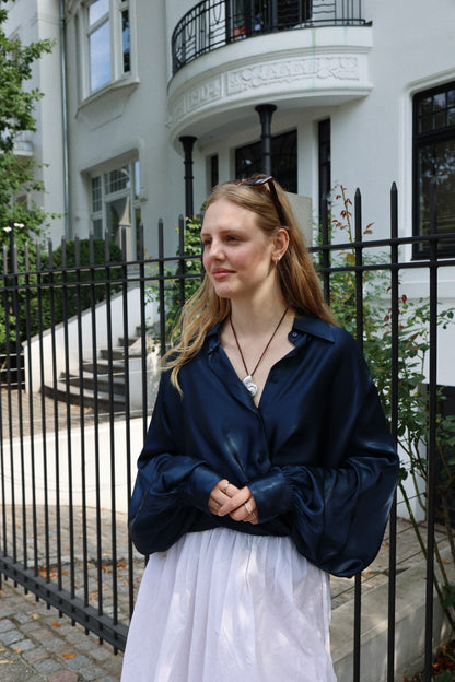 Eine junge Frau mit langen blonden Haaren, die die elegante Satinbluse Antonia Shiny Navy und einen weißen, übergroßen Rock trägt, steht vor einem schwarzen Eisenzaun und einem großen weißen Gebäude mit gewölbten Fenstern und schaut lächelnd zur Seite.