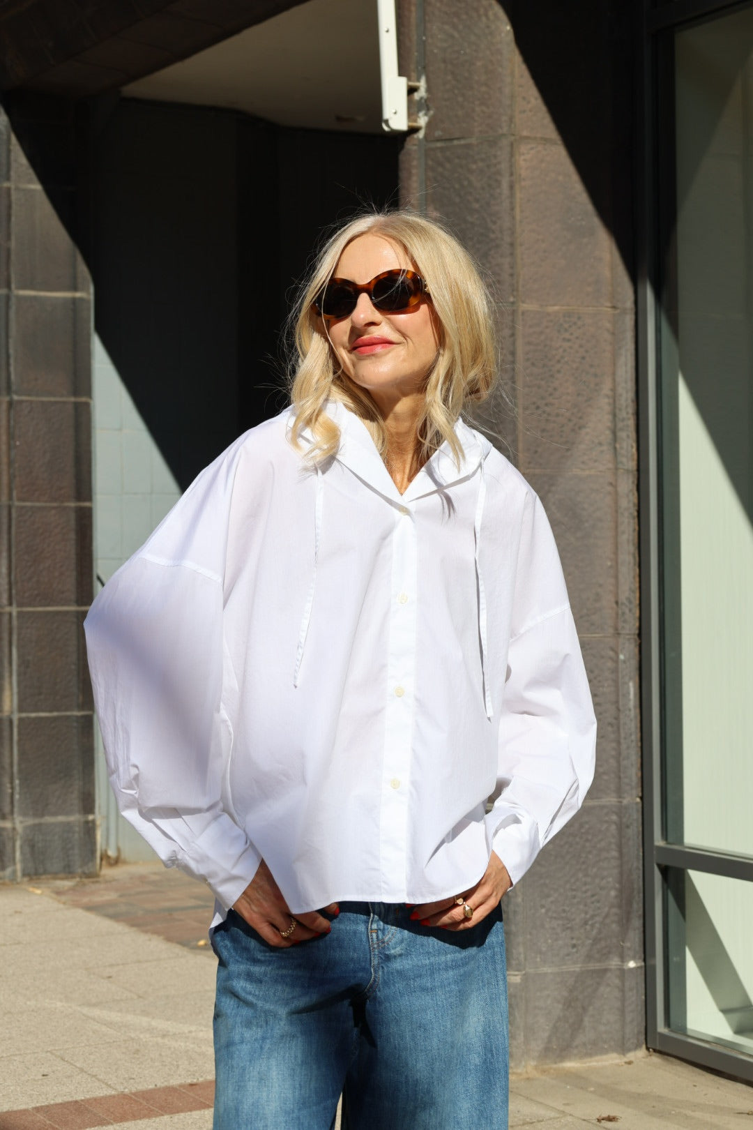 Eine Frau mit langen blonden Haaren und Sonnenbrille, die einen weißen Bluse Hoodie und blaue Jeans trägt, steht draußen im Sonnenlicht und lächelt mit den Händen in den Taschen.