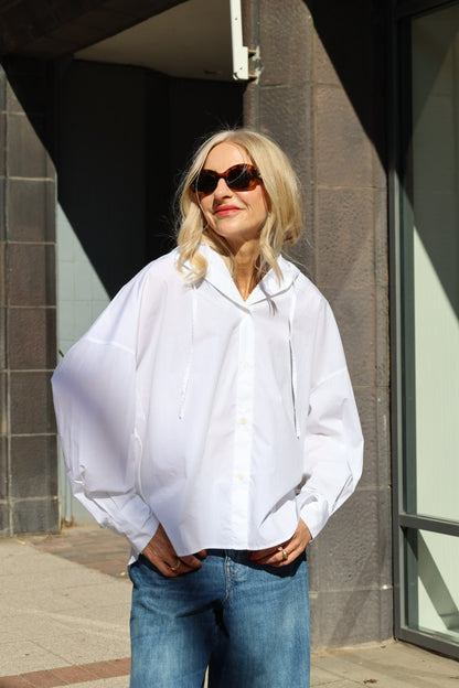 Eine Frau mit langen blonden Haaren und Sonnenbrille, die einen weißen Bluse Hoodie und blaue Jeans trägt, steht draußen im Sonnenlicht und lächelt mit den Händen in den Taschen.