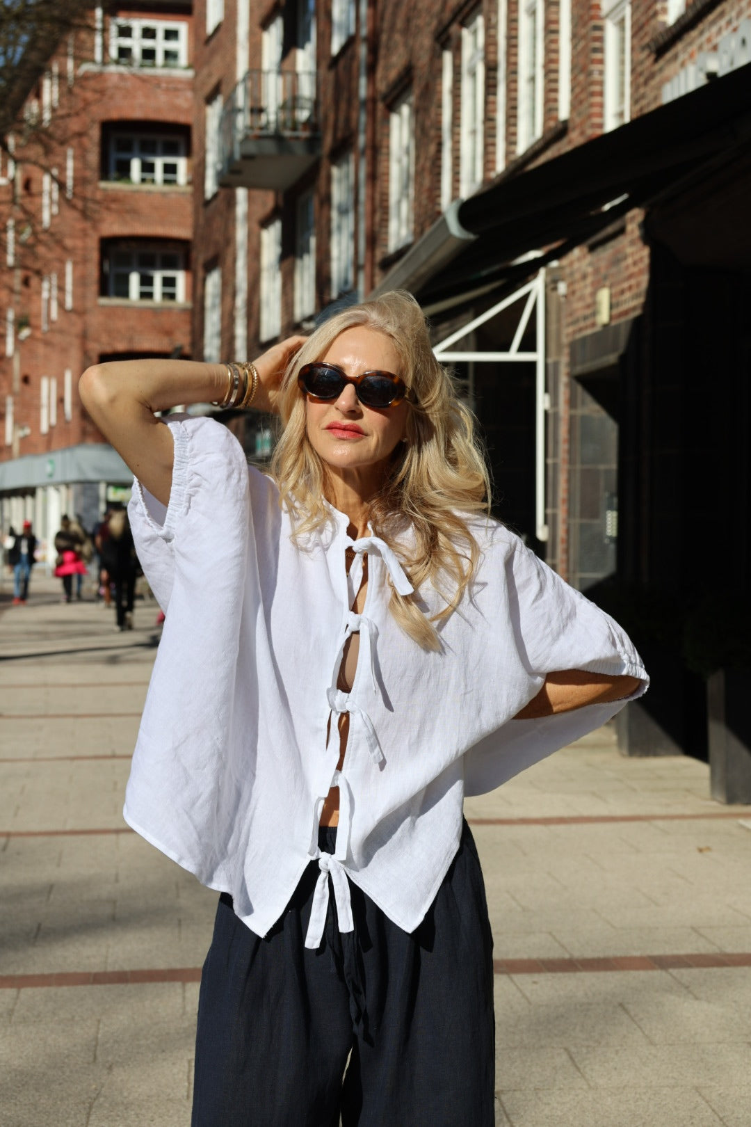 Eine Frau mit langen blonden Haaren und Sonnenbrille steht selbstbewusst in dunklen Hosen und der Bluse Cowboy Bow auf einer sonnigen Stadtstraße und zeigt ihre Liebe zur nachhaltigen Mode.