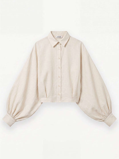 Die Bluse „Antonia Short Flanell Offwhite“ – eine beigefarbene, kurz geschnittene Bluse aus hochwertigem Flanell, hergestellt in Portugal – verfügt über einen Kragen, eine Knopfleiste vorne und auffällige Ballonärmel, die an den Manschetten gerafft sind. Das Kleidungsstück wird flach auf einem weißen Hintergrund präsentiert.