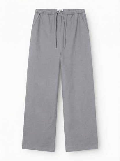 Die Hose „Flanell Grau“ ist eine hellgraue Hose mit weitem Bein, die über einen elastischen Bund, einen Kordelzug und Seitentaschen verfügt. Sie besteht aus 100 % Baumwolle und wird vor einem schlichten weißen Hintergrund gezeigt.