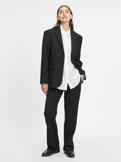 Eine Person steht vor einem schlichten weißen Hintergrund, trägt die Hose Karoline Black, ein weißes Button-up-Hemd und schwarze Schuhe und blickt in einer entspannten Pose leicht nach oben, eine Hand in der Tasche.