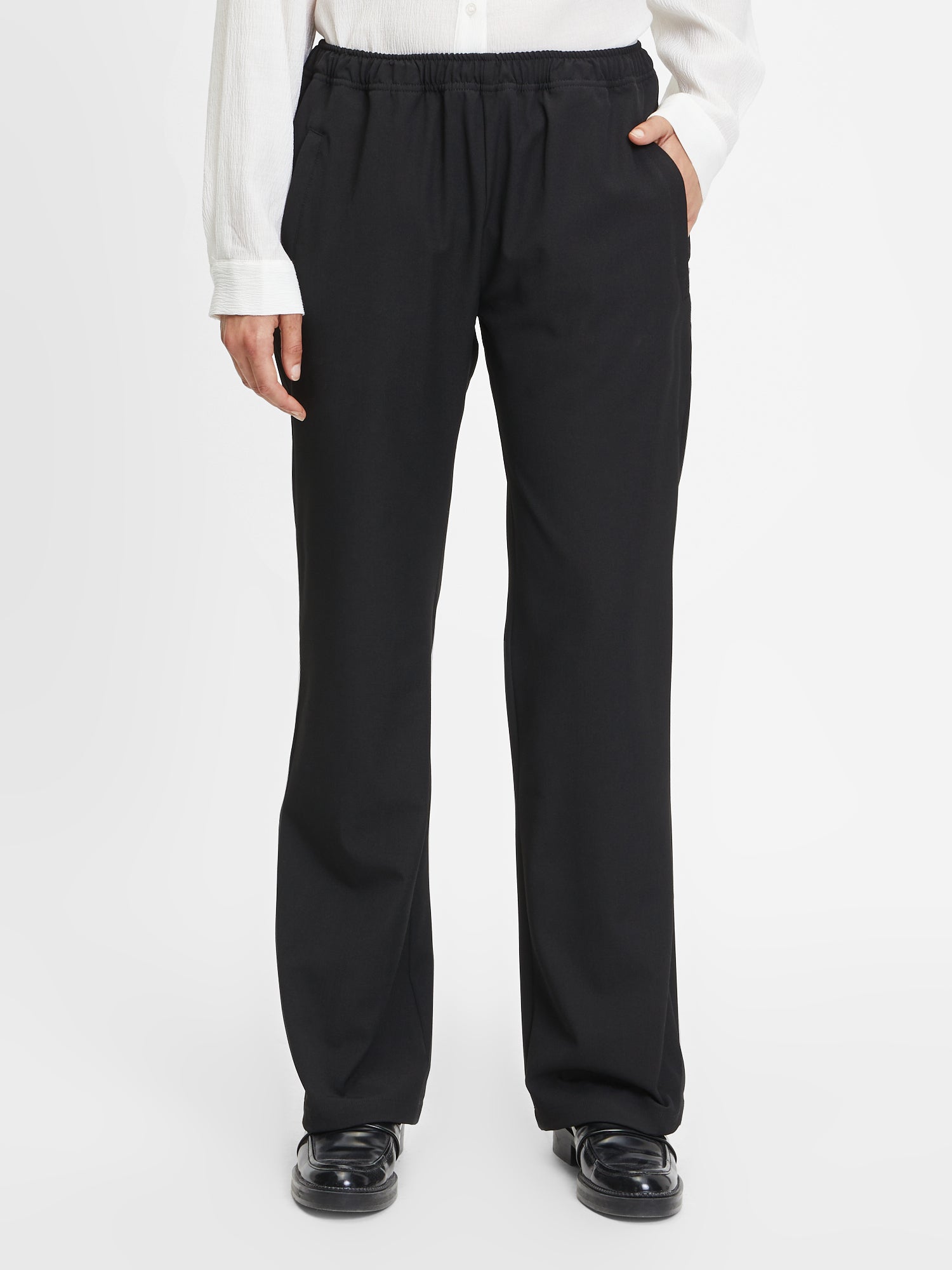 Eine Person, die ein weißes langärmeliges Hemd und eine Hose Karoline Black mit den Händen in den Taschen trägt, steht vor einem schlichten weißen Hintergrund, der nur die untere Hälfte dieses Businessoutfits Damen zeigt.