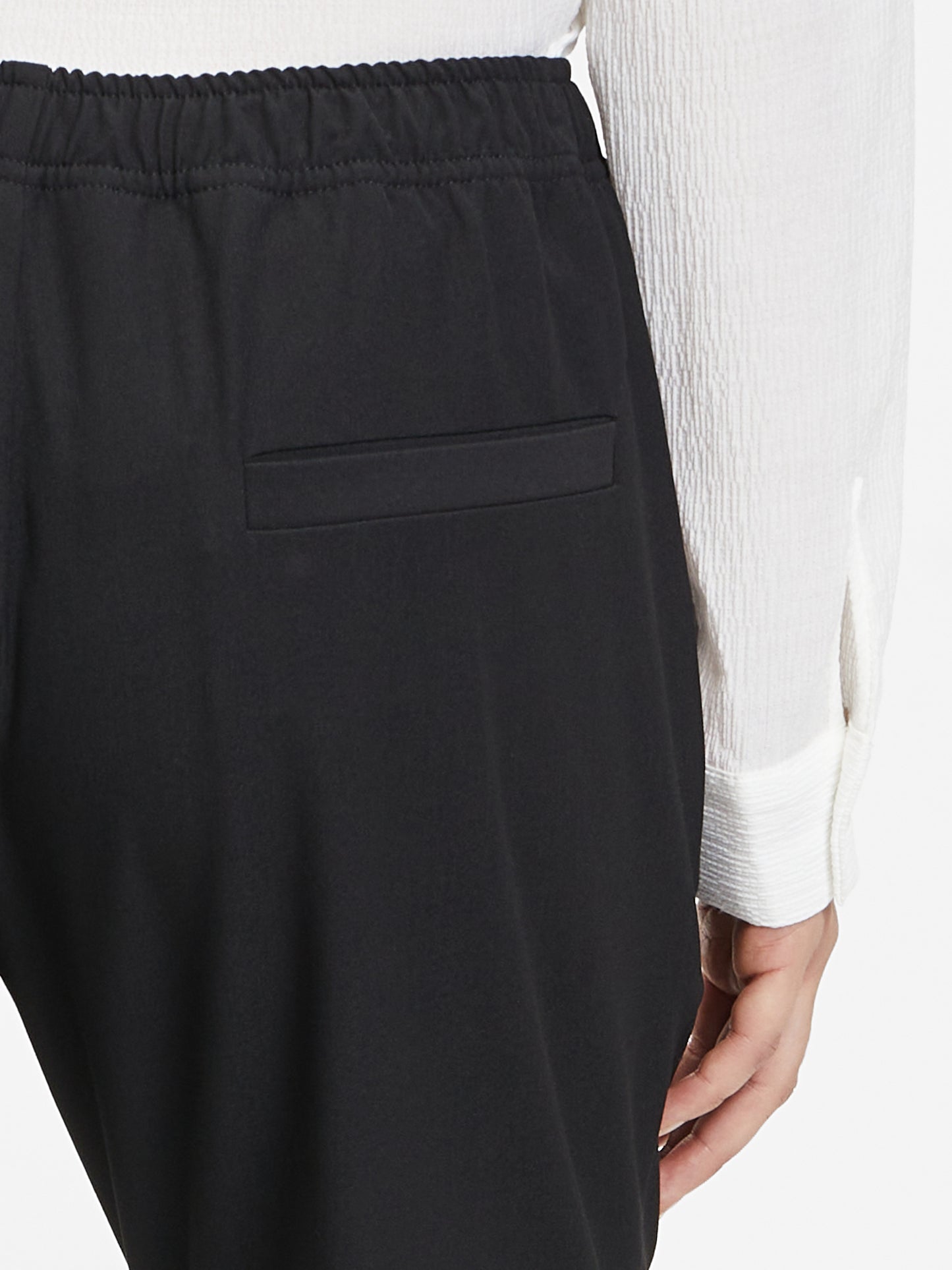Man sieht eine Person von hinten, die die Hose Karoline Black mit Gummibund und ein weißes langärmeliges Hemd trägt; nur der untere Rücken und eine Hand sind zu sehen.