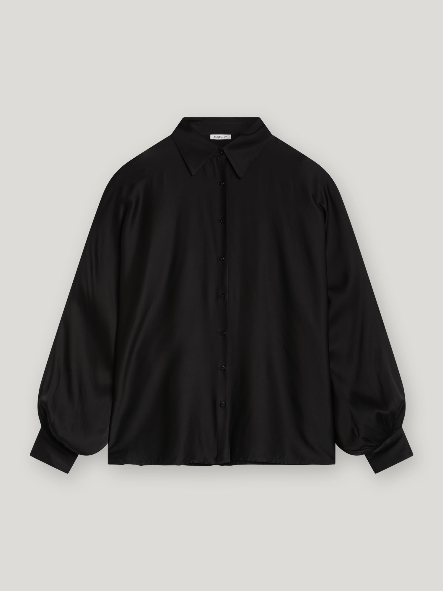 Die Bluse Antonia Black ist eine locker sitzende, langärmelige Bluse mit spitzem Kragen und breiten Manschetten, die auf einem unifarbenen hellgrauen Hintergrund abgebildet ist.