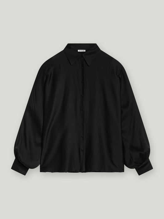 Die Bluse Antonia Black ist eine locker sitzende, langärmelige Bluse mit spitzem Kragen und breiten Manschetten, die auf einem unifarbenen hellgrauen Hintergrund abgebildet ist.