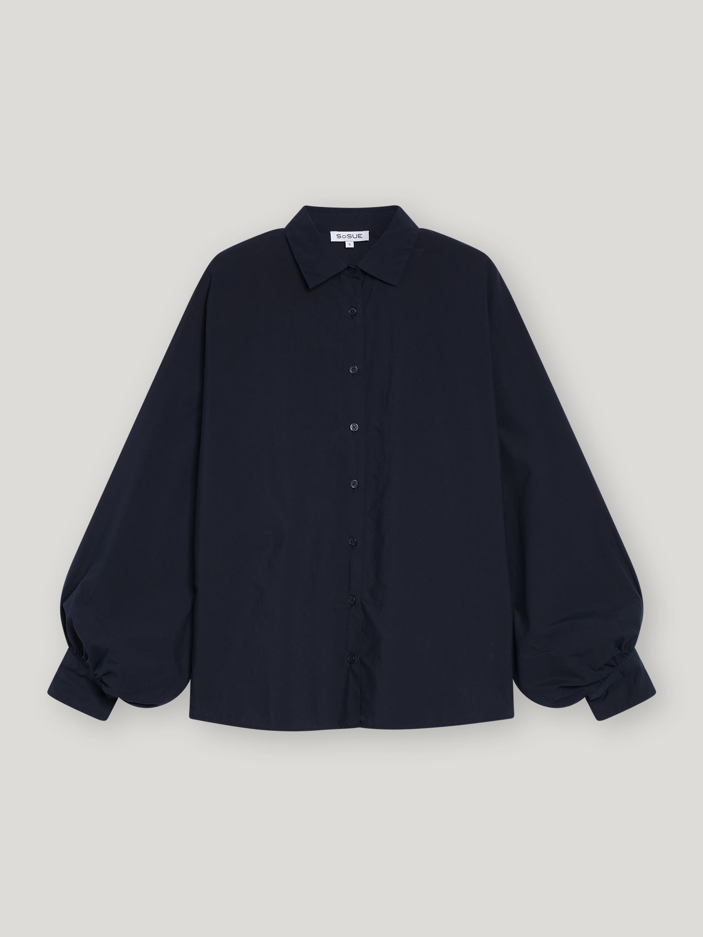 Die Bluse Antonia Slim Dark Blue, eine schmal geschnittene Damenbluse mit langen Ärmeln, Kragen und voluminös gerafften Manschetten, präsentiert sich elegant vor einem schlichten hellgrauen Hintergrund.