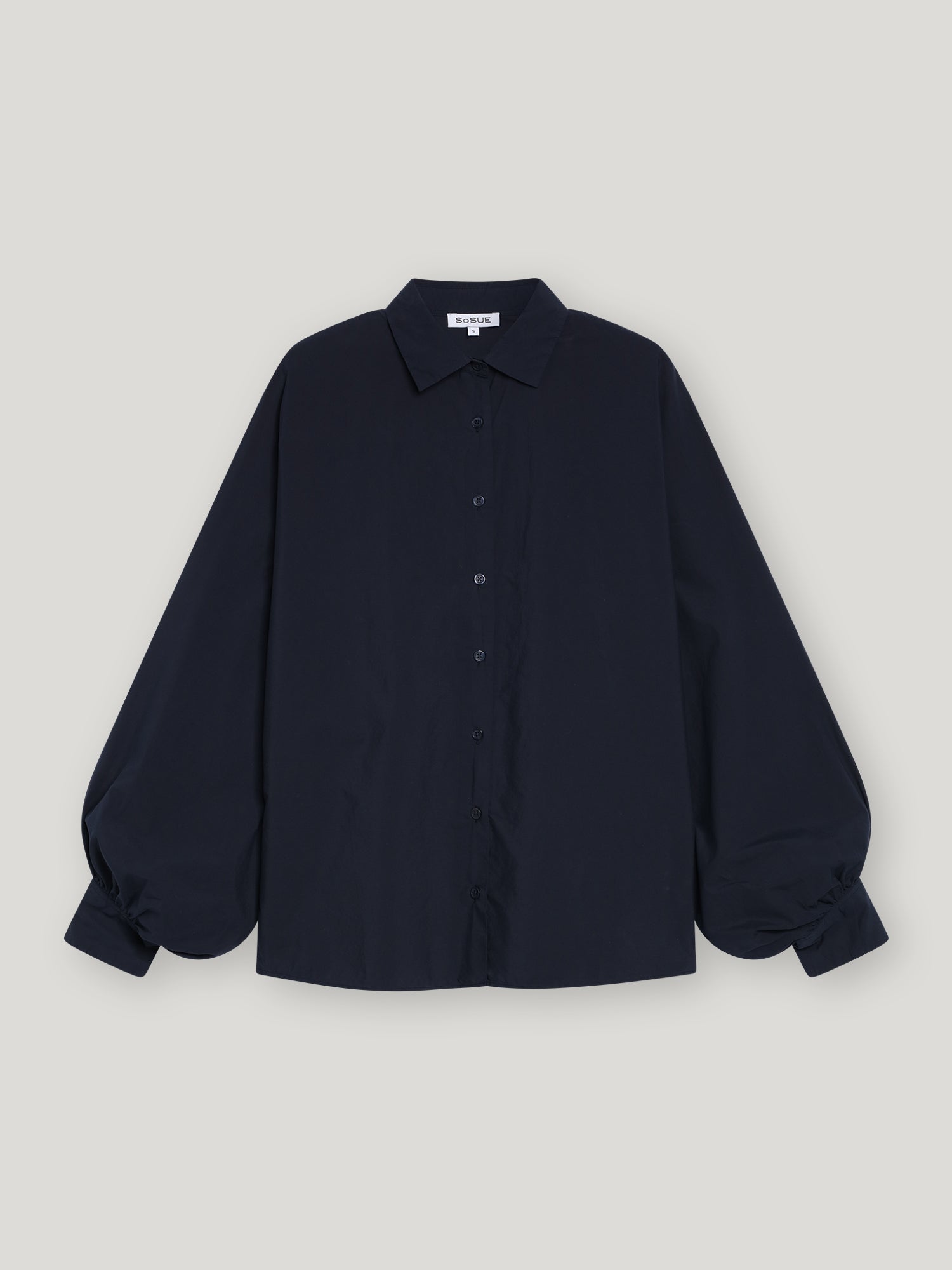 Die Bluse Antonia Slim Dark Blue, eine schmal geschnittene Damenbluse mit langen Ärmeln, Kragen und voluminös gerafften Manschetten, präsentiert sich elegant vor einem schlichten hellgrauen Hintergrund.