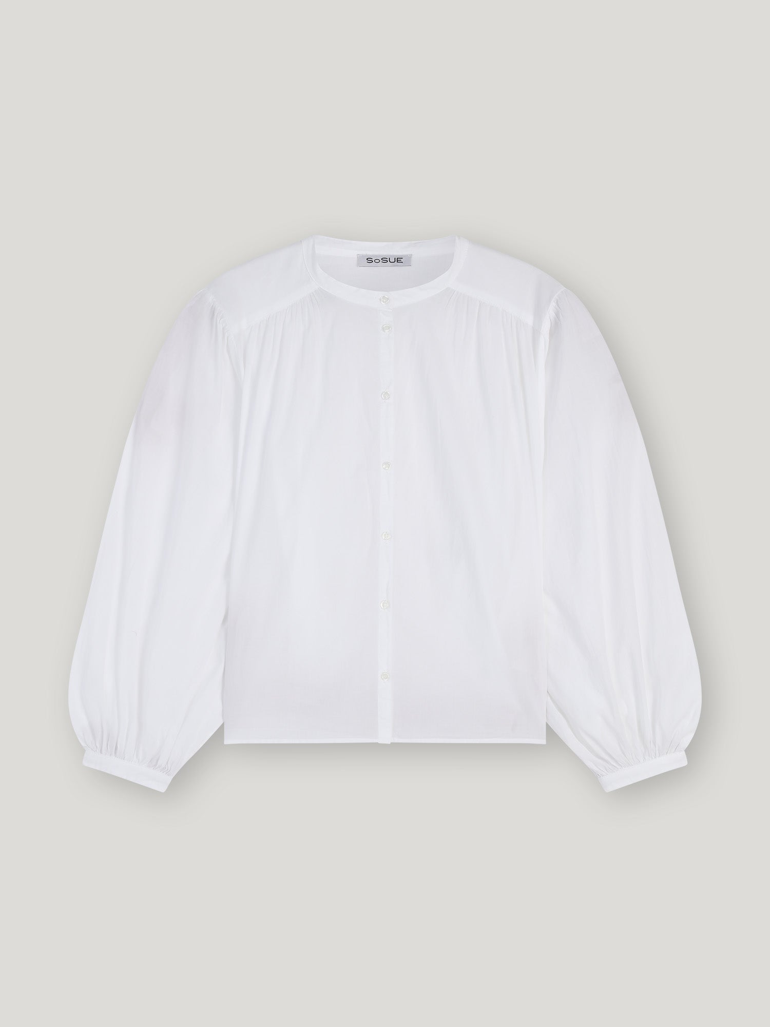 Die Bluse Bella White hat einen Rundhalsausschnitt, eine geknöpfte Vorderseite, geraffte Schultern und Puffärmel mit geknöpften Manschetten in einer übergroßen Silhouette, die sich vor einem hellgrauen Hintergrund präsentiert.