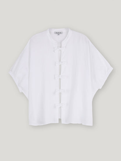 Die Bluse Cowboy Bow ist eine locker sitzende weiße Leinenbluse für Damen mit kurzen Ärmeln, Mandarinkragen und fünf traditionellen Knotenverschlüssen auf der Vorderseite. Auf hellgrauem Grund ist sie die ideale Wahl für Fans der nachhaltigen Mode.