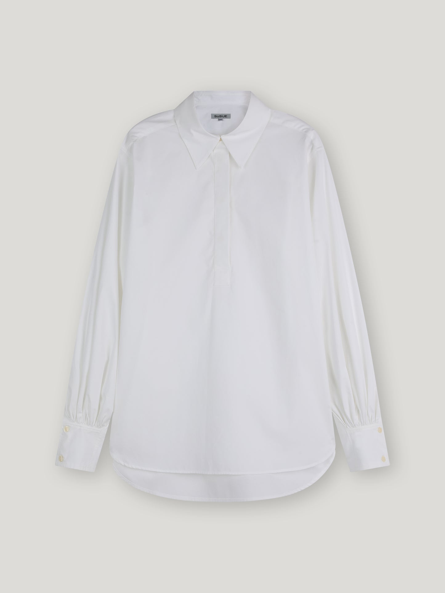 Die Bluse Rini White ist eine übergroße, langärmelige Popeline-Baumwollbluse mit spitzem Kragen und geknöpften Manschetten, die vor einem schlichten, hellen Hintergrund gezeigt wird.