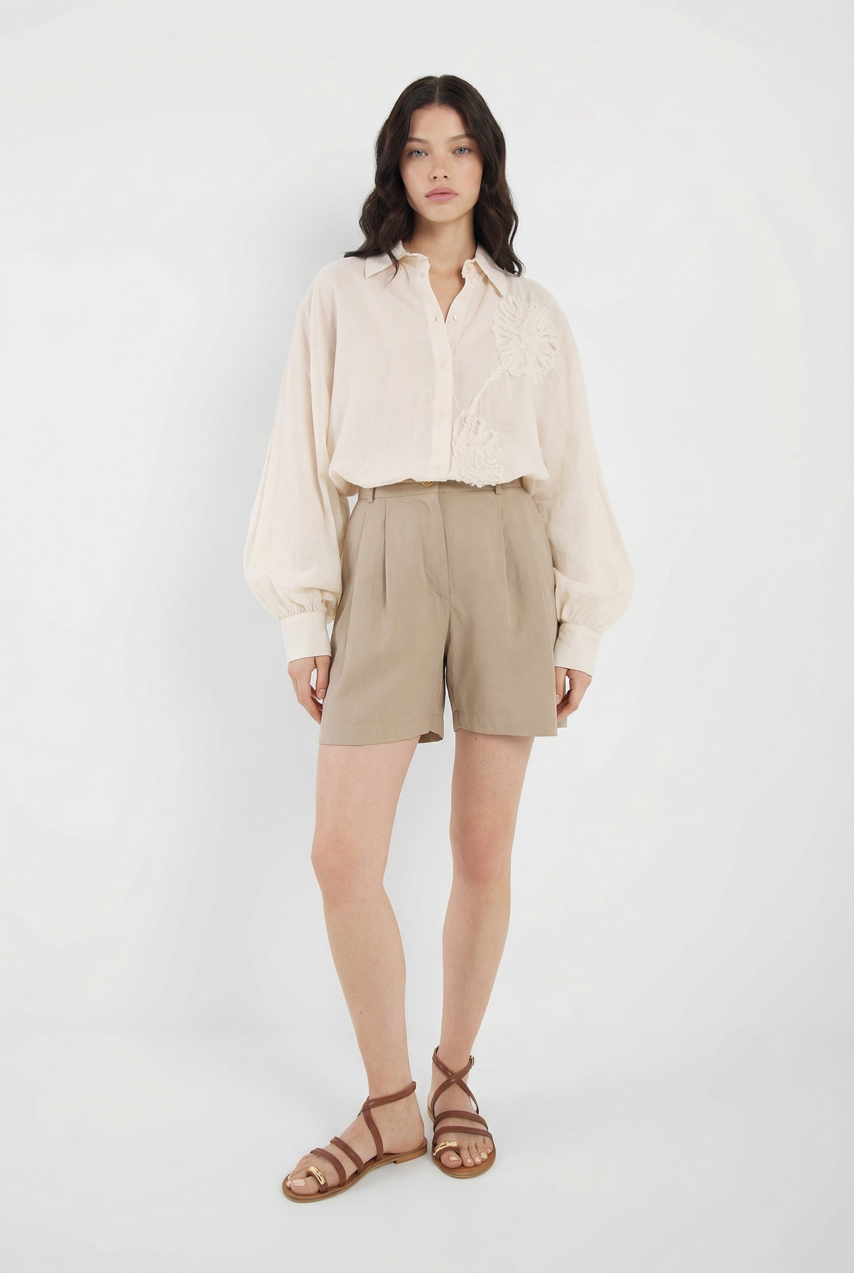Eine Frau mit langen dunklen Haaren und neutralem Gesichtsausdruck steht vor einem weißen Hintergrund. Sie trägt die Bluse Antonia Crafted Panna, hellbraune, hoch taillierte Shorts und braune Riemchensandalen.
