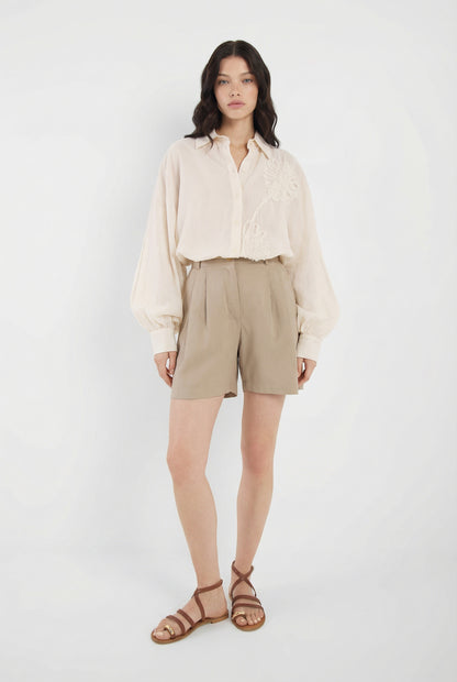 Eine Frau mit langen dunklen Haaren und neutralem Gesichtsausdruck steht vor einem weißen Hintergrund. Sie trägt die Bluse Antonia Crafted Panna, hellbraune, hoch taillierte Shorts und braune Riemchensandalen.