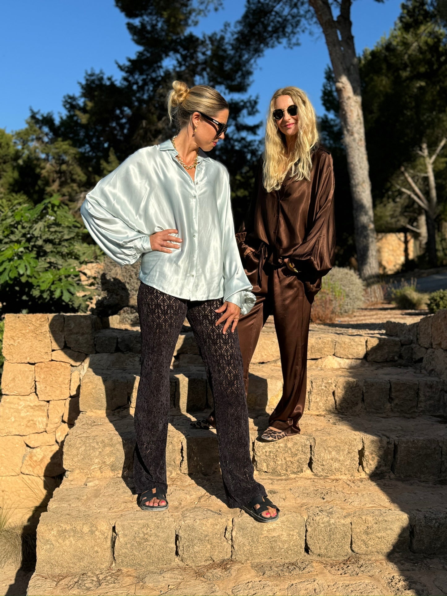 Zwei Frauen stehen im Freien auf steinernen Stufen, beide in übergroßen Satinhemden - eine trägt das Bluse Antonia Shiny Slim IceGrey, die andere dunkelbraun. Stilvolle Sonnenbrillen und Posen unterstreichen die Eleganz dieser Satin-Looks.