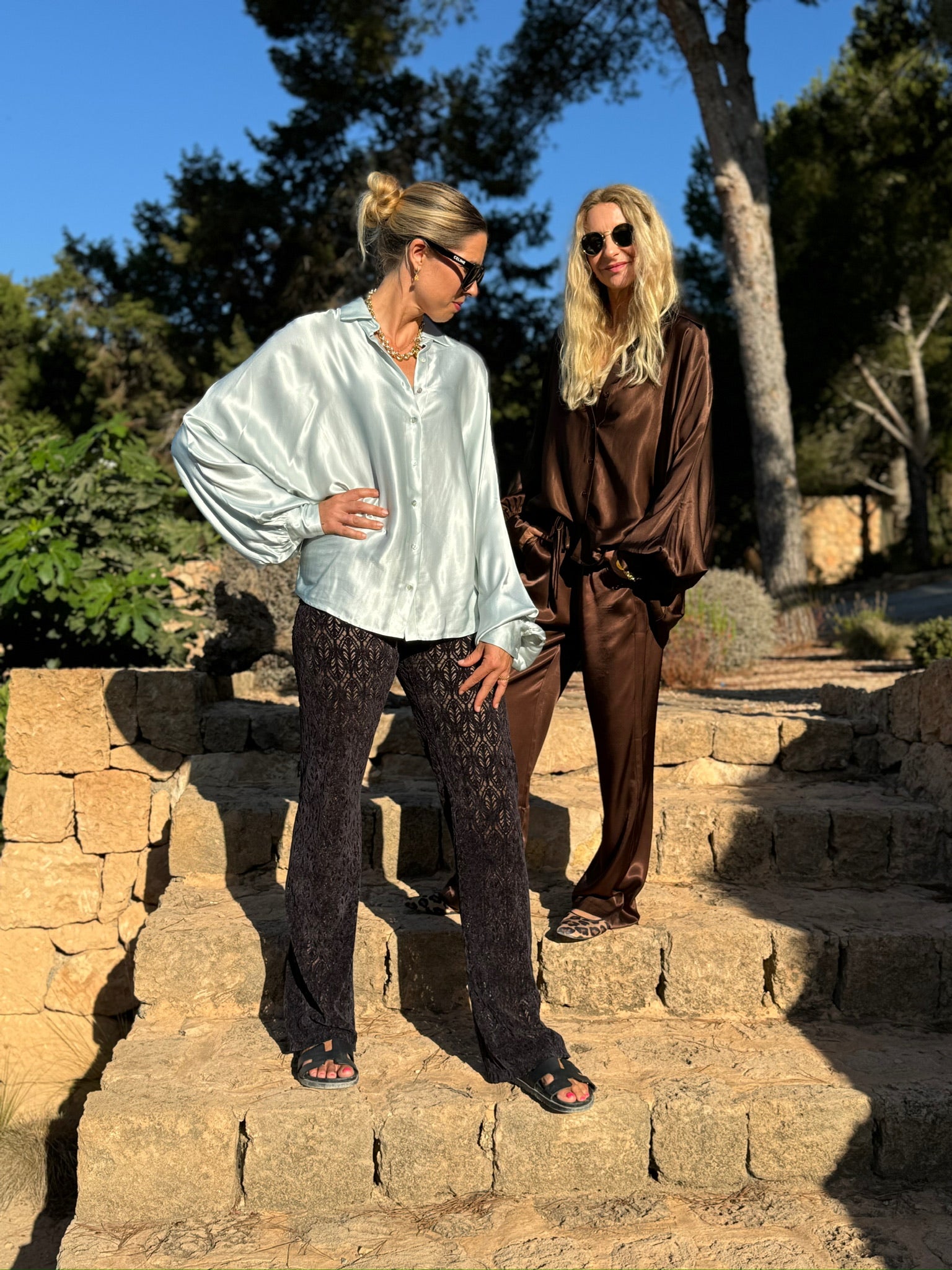 Zwei Frauen stehen im Freien auf steinernen Stufen, beide in übergroßen Satinhemden - eine trägt das Bluse Antonia Shiny Slim IceGrey, die andere dunkelbraun. Stilvolle Sonnenbrillen und Posen unterstreichen die Eleganz dieser Satin-Looks.