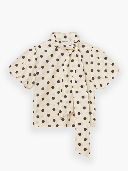 Bluse Bow Dots Ecru Schwarz: Ecru-Bluse mit großen schwarzen Tupfen, kurzen Ärmeln, hohem Halsausschnitt und einer langen, lockeren seitlichen Schalbindung.