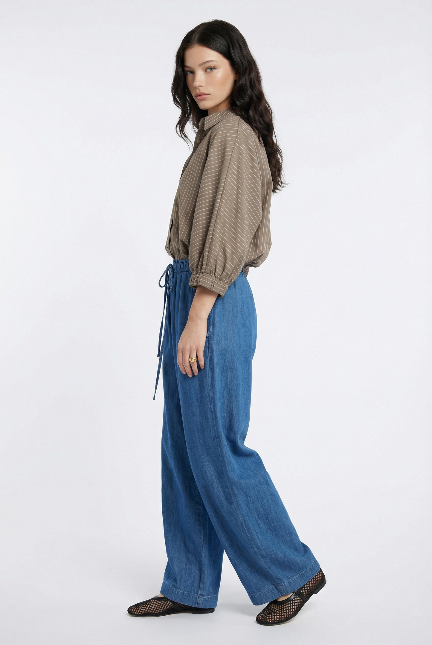 Eine Frau mit langen dunklen Haaren steht im Profil vor einem weißen Hintergrund und trägt die Bluse Antonia Short Sleeve Taupe Stripes mit einer blauen Hose mit weitem Bein und schwarzen gewebten Flats.