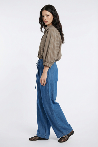 Eine Frau mit langen dunklen Haaren steht im Profil vor einem weißen Hintergrund und trägt die Bluse Antonia Short Sleeve Taupe Stripes mit einer blauen Hose mit weitem Bein und schwarzen gewebten Flats.