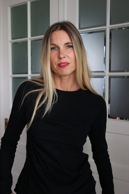 Eine Frau mit langen blonden Haaren trägt das Shirt Gathering Black und steht in einem Innenraum vor einer weißen Glastür. Sie blickt mit neutraler Miene in die Kamera und reflektiert nachhaltige Frauenmode.