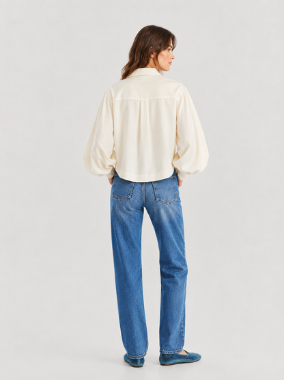 Eine Frau steht mit dem Rücken zur Kamera und trägt die langärmelige Bluse „Antonia Short Flanell Offwhite“, kombiniert mit einer blauen Jeans und türkisfarbenen Ballerinas, vor einem hellgrauen Hintergrund. Diese hochwertige Flanellbluse wird in Portugal hergestellt und besticht sowohl durch ihre Eleganz als auch durch ihre außergewöhnliche Verarbeitungsqualität.