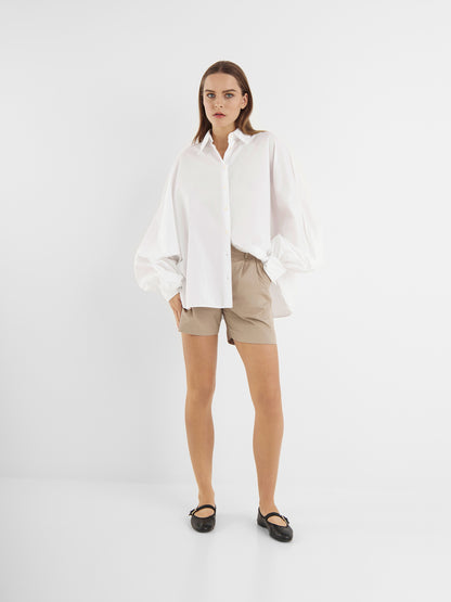 Eine Frau steht vor einem weißen Hintergrund und trägt die Bluse Antonia White, eine lockere Baumwollbluse mit weiten Ärmeln, kombiniert mit beigen Shorts und schwarzen Ballerinas. Sie blickt mit neutralem Blick nach vorne.