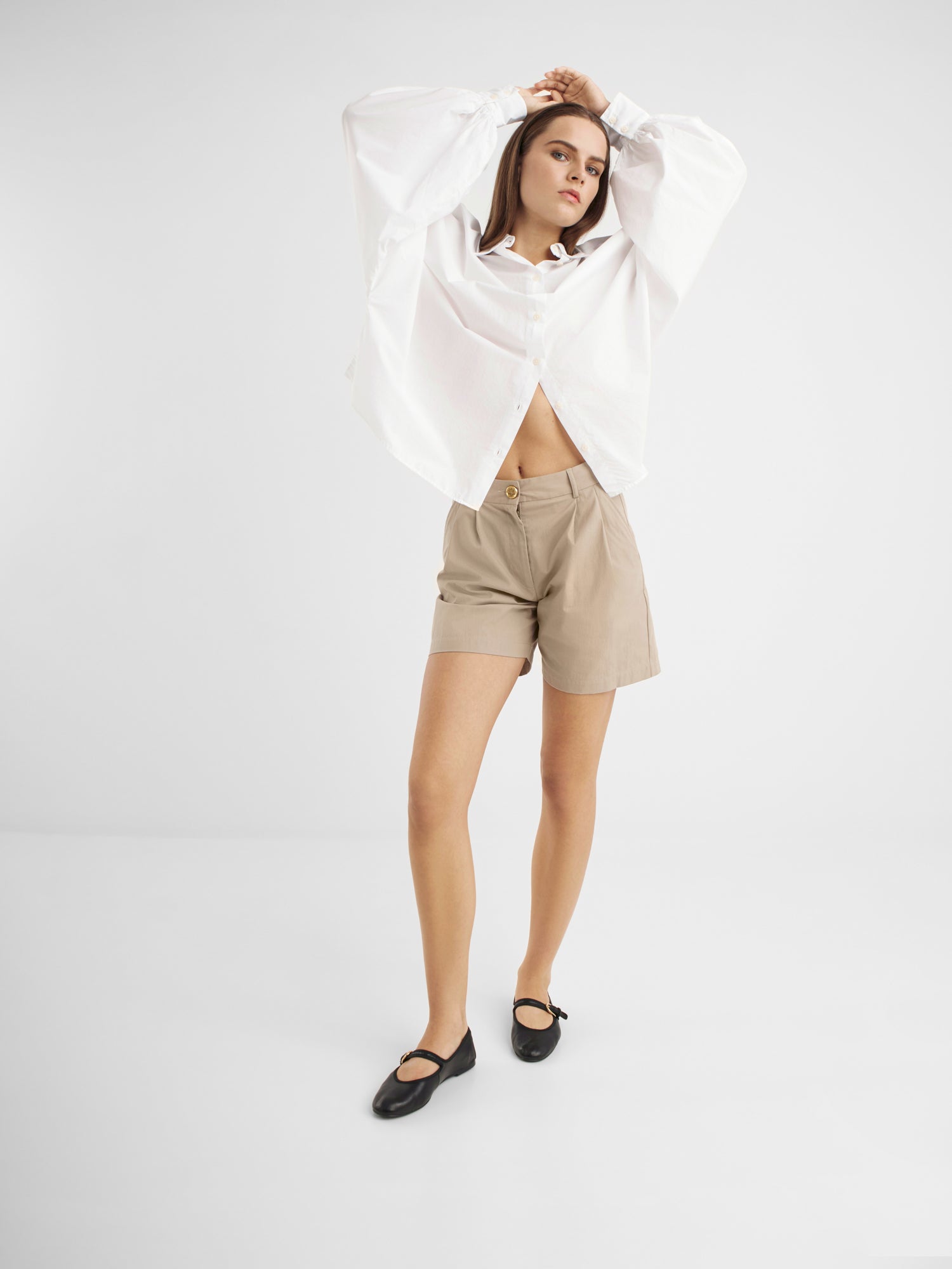 Eine Frau posiert vor einem schlichten weißen Hintergrund. Sie trägt eine beigefarbene, hoch taillierte Shorts Safari, ein weißes, übergroßes Hemd und schwarze flache Schuhe. Sie hebt einen Arm und blickt nachdenklich zur Seite.