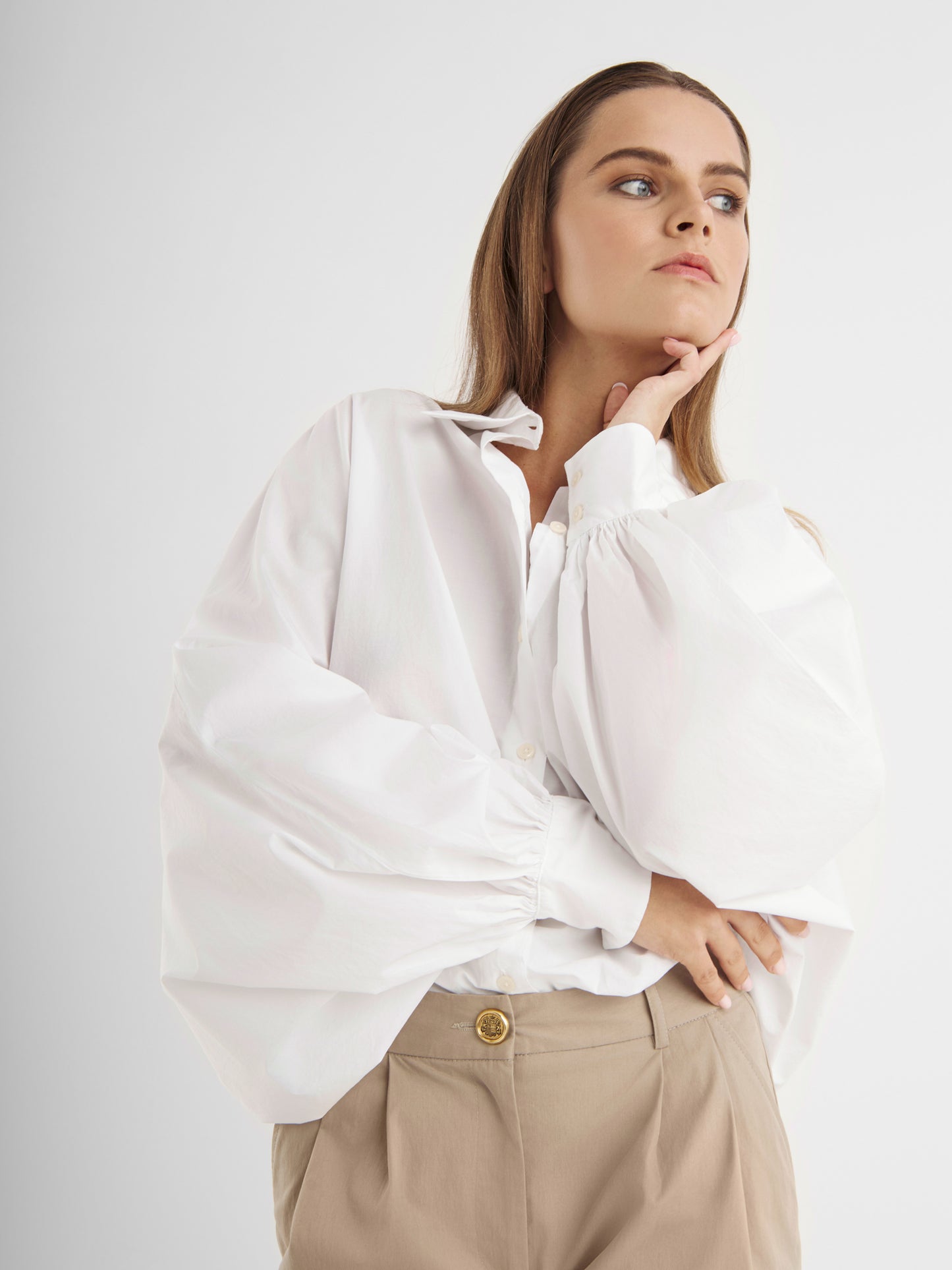Eine Frau mit langen braunen Haaren trägt die Bluse Antonia White - eine elegante weiße Baumwollbluse mit Puffärmeln - und eine beigefarbene Hose. Sie stützt ihr Kinn auf ihre Hand und blickt nachdenklich zur Seite vor einem schlichten Hintergrund.