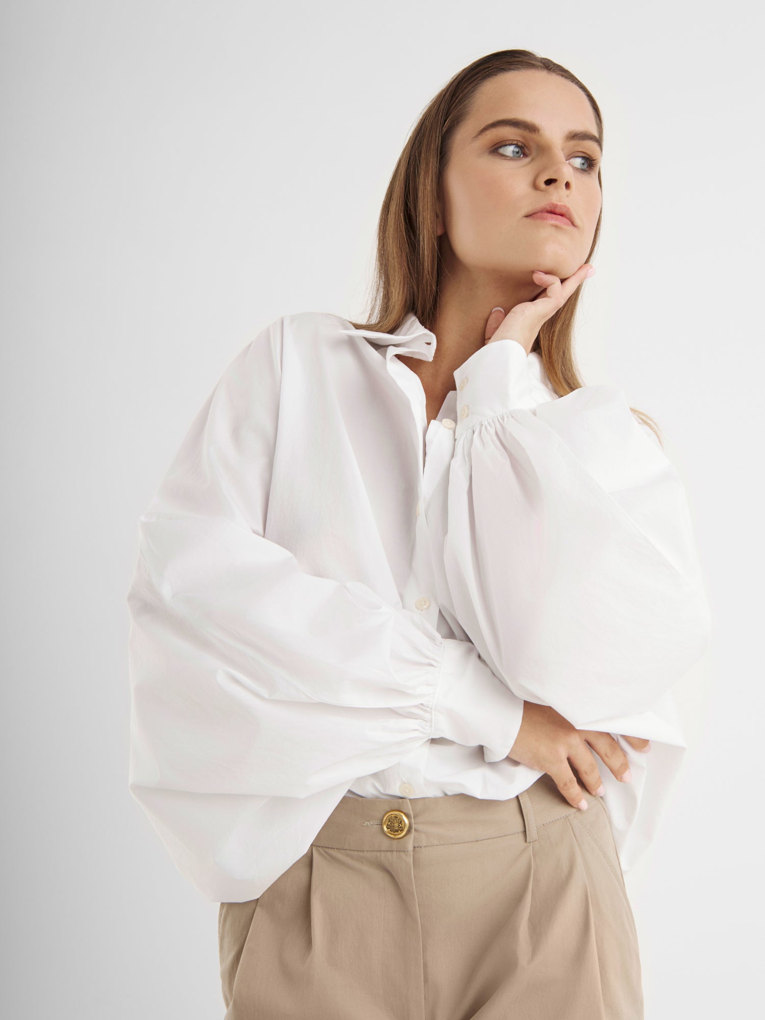 Eine Frau mit langen braunen Haaren trägt die Bluse Antonia White - eine elegante weiße Baumwollbluse mit Puffärmeln - und eine beigefarbene Hose. Sie stützt ihr Kinn auf ihre Hand und blickt nachdenklich zur Seite vor einem schlichten Hintergrund.