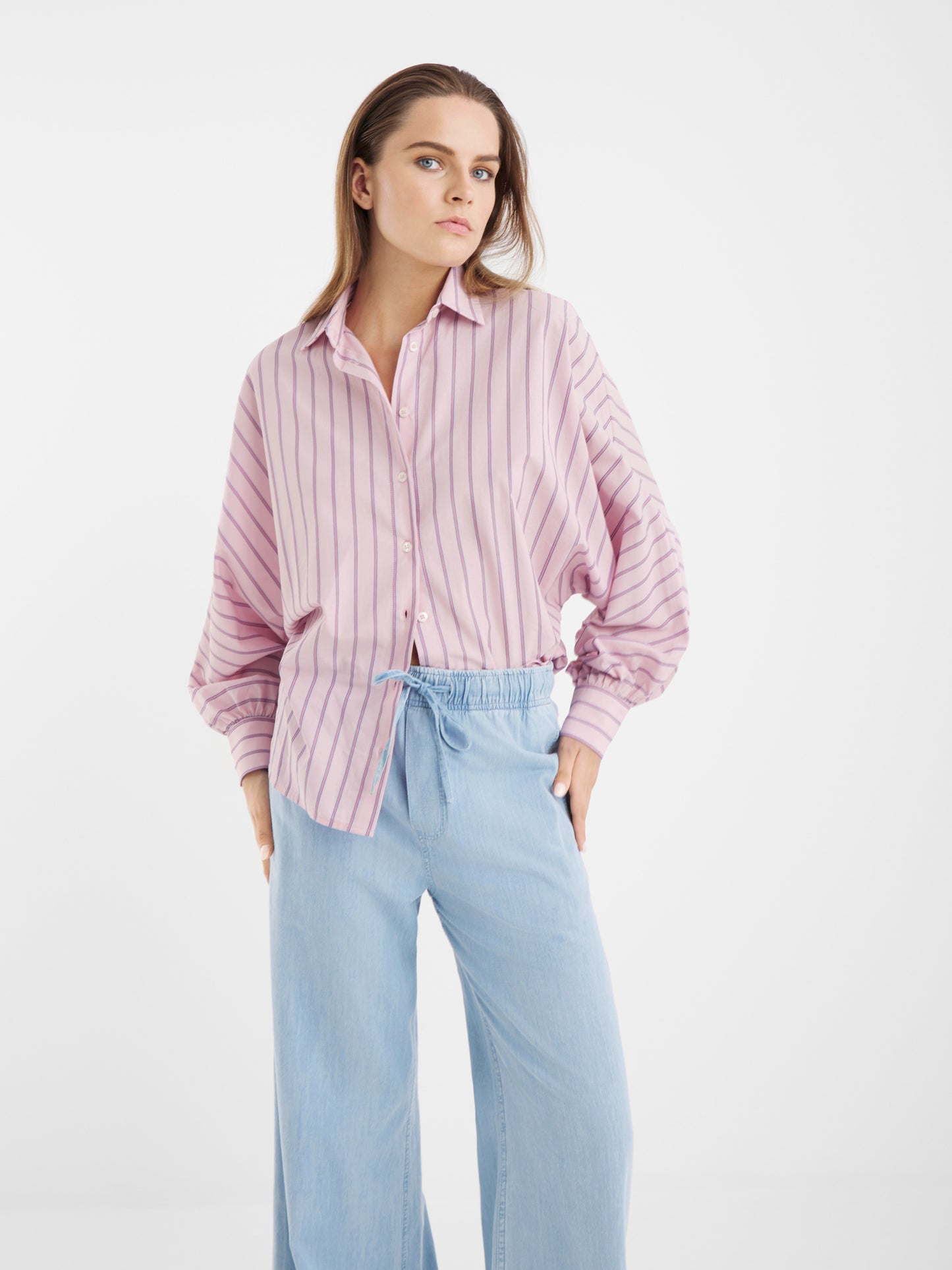 Eine Frau mit glattem, schulterlangem Haar und neutralem Gesichtsausdruck trägt das Button-up-Hemd Antonia Slim Stripes Rose vor einem schlichten weißen Hintergrund, gepaart mit einer hellblauen Hose mit weitem Bein und elastischem Bund.
