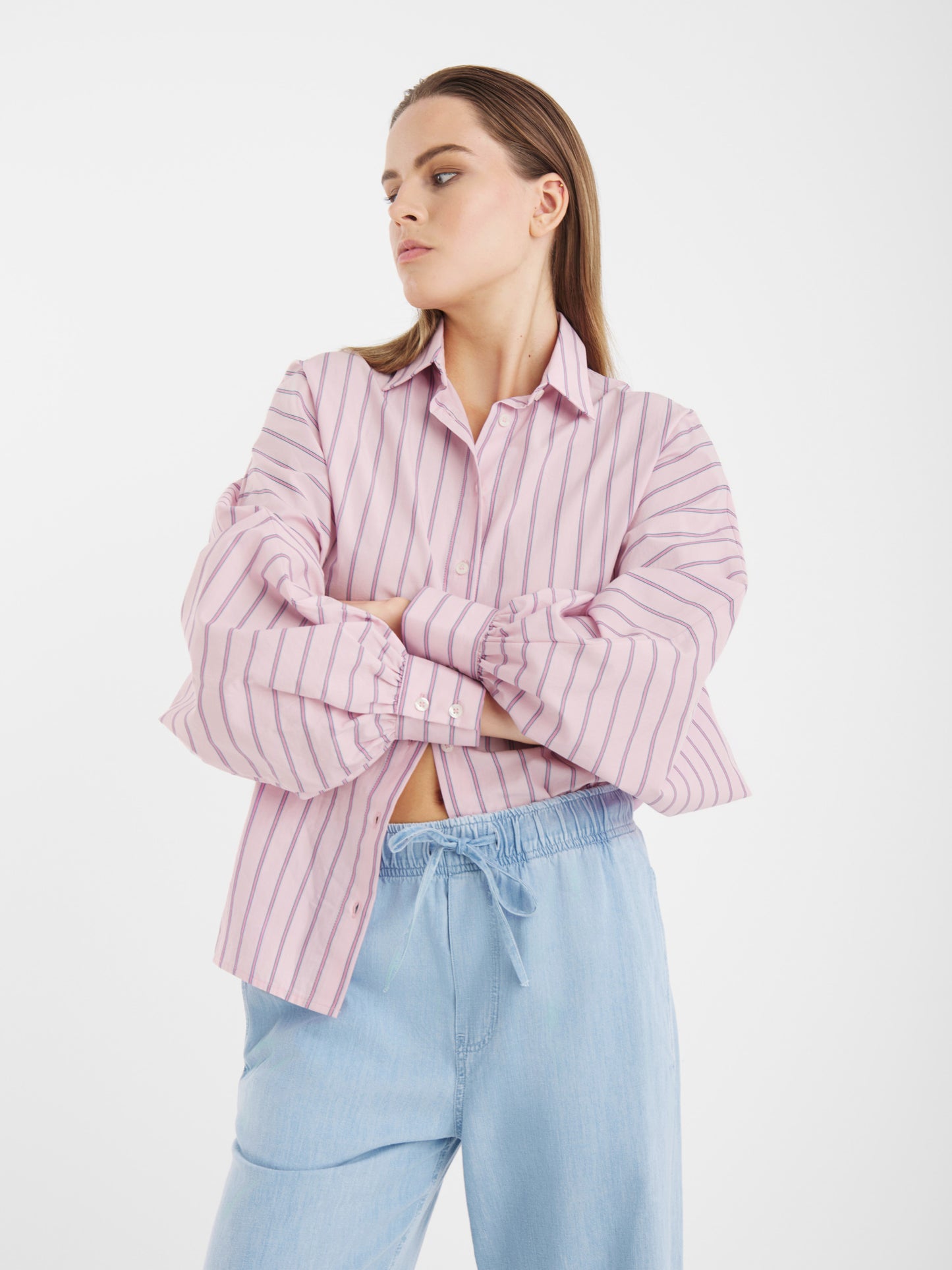 Eine Frau trägt die SOSUE Bluse Antonia Slim Rose Stripes mit weiten Ärmeln und einer hellblauen Hose mit Kordelzug. Sie steht vor einem schlichten weißen Hintergrund und blickt mit verschränkten Armen zur Seite.