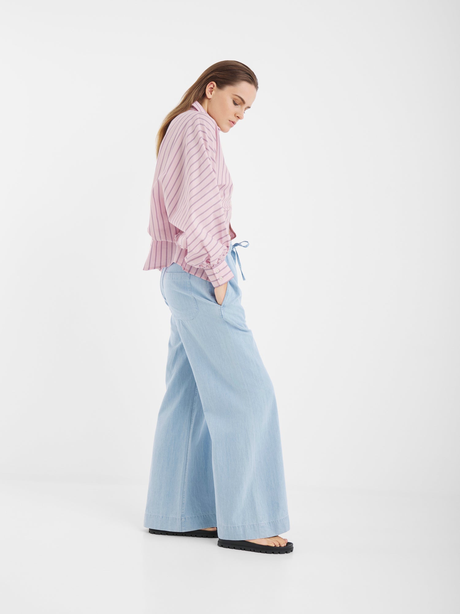 Frau im Profil vor weißem Hintergrund trägt Anna Pants Light Denim, eine hellrosa gestreifte Bluse mit Ballonärmeln und schwarze Schuhe. Sie steht mit den Händen in den Taschen und blickt nach unten.