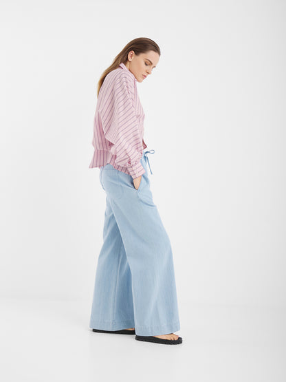Frau im Profil vor weißem Hintergrund trägt Anna Pants Light Denim, eine hellrosa gestreifte Bluse mit Ballonärmeln und schwarze Schuhe. Sie steht mit den Händen in den Taschen und blickt nach unten.