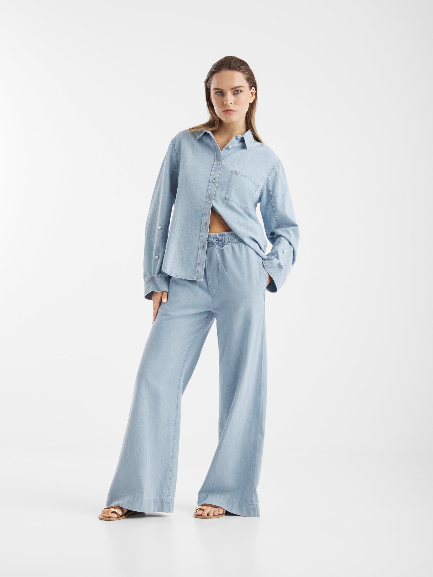 Eine Frau blickt vor einem weißen Hintergrund nach vorne und trägt Anna Pants Light Denim - eine locker sitzende Hose mit weitem Bein, gepaart mit einem passenden hellblauen Jeanshemd und Sandalen. Eine Hand steckt in ihrer Tasche.