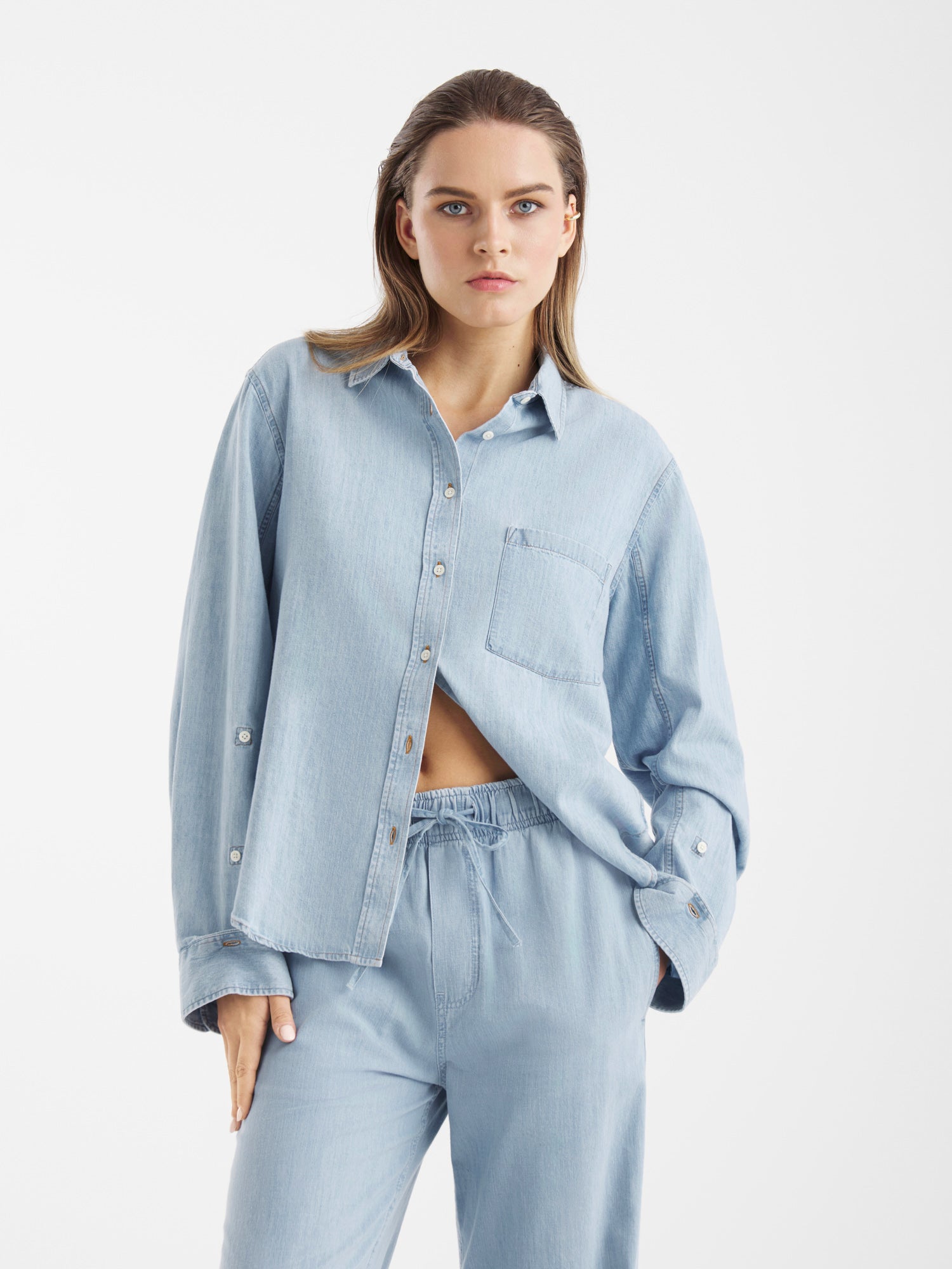 Eine Frau steht vor einem weißen Hintergrund und trägt eine Blouse Classic Denim in Hellblau mit einer Brusttasche und eine dazu passende lockere Hose mit Kordelzug. Ihre linke Hand ruht in ihrer Tasche, während sie selbstbewusst in die Kamera blickt.