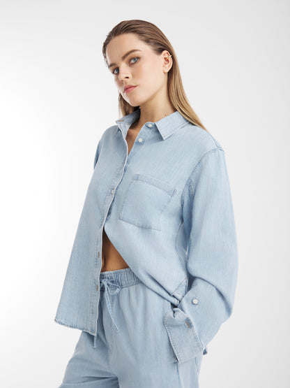 Eine Frau, die die Blouse Classic Denim in Hellblau und die dazu passende, tailliert geschnittene Hose trägt, steht vor einem weißen Hintergrund und blickt selbstbewusst in die Kamera, eine Hand in der Tasche.