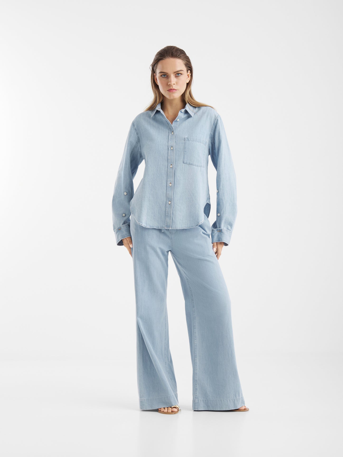 Eine Frau steht mit dem Gesicht nach vorne und trägt die Bluse Classic Denim in Hellblau mit tailliertem Schnitt und eine passende Hose mit weitem Bein, beide aus Denim Chambray. Der Hintergrund ist einfarbig weiß.