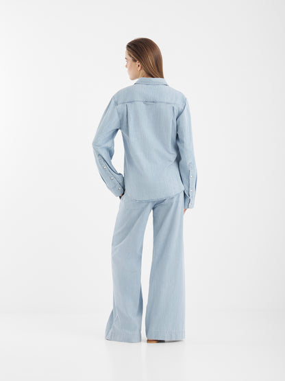 Eine Frau blickt von sich weg und trägt die Blouse Classic Denim in Hellblau mit einer dazu passenden, locker sitzenden Jeans mit weitem Bein vor einem schlichten weißen Hintergrund.