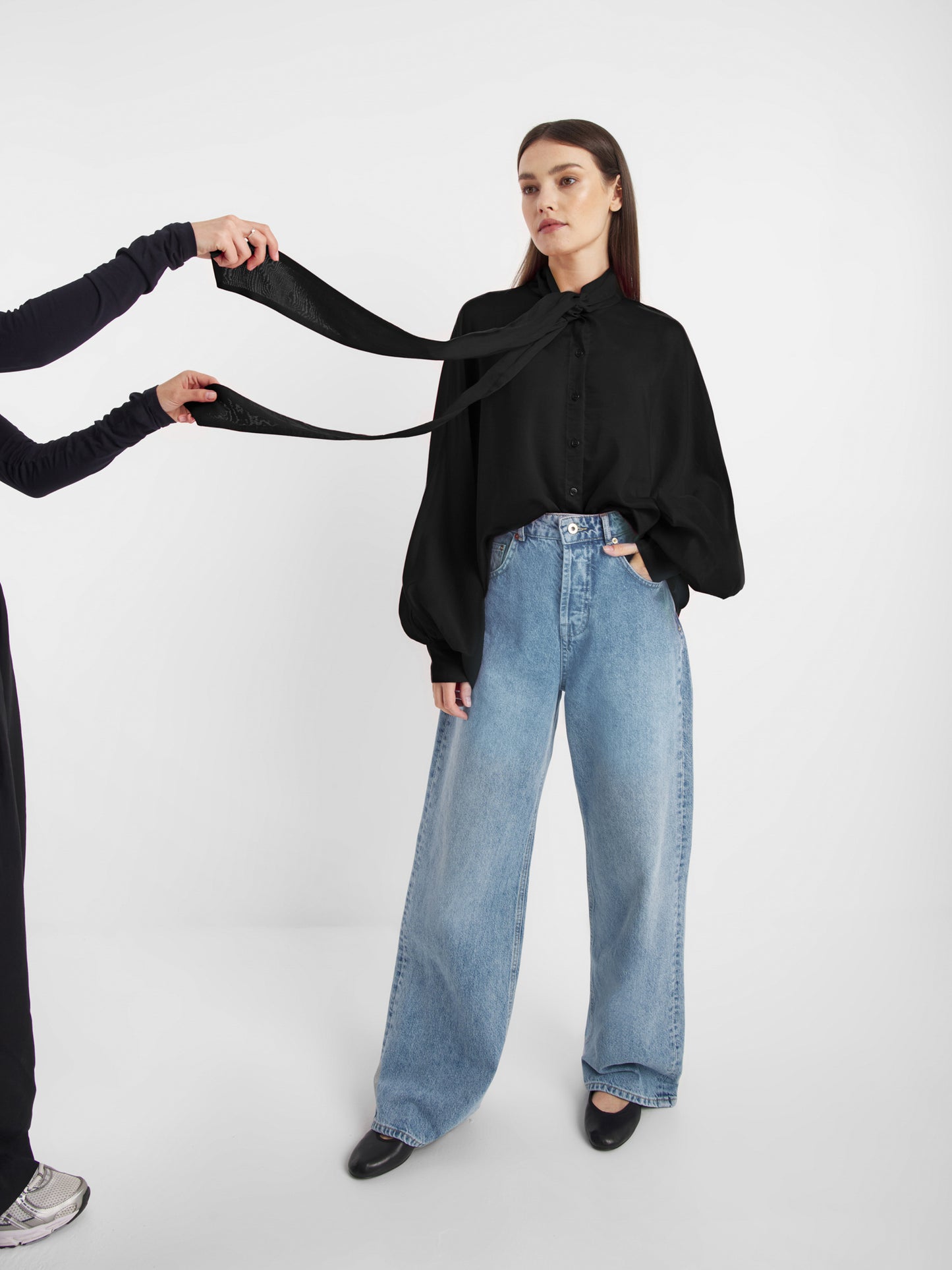 Eine Frau in einer schwarzen SOSUE-Schluppenbluse und weit geschnittenen Bluejeans steht da, während eine andere Person, meist außerhalb des Bildausschnitts, ihre lange schwarze Antonia Bow-Krawatte vor einem schlichten weißen Hintergrund bindet.