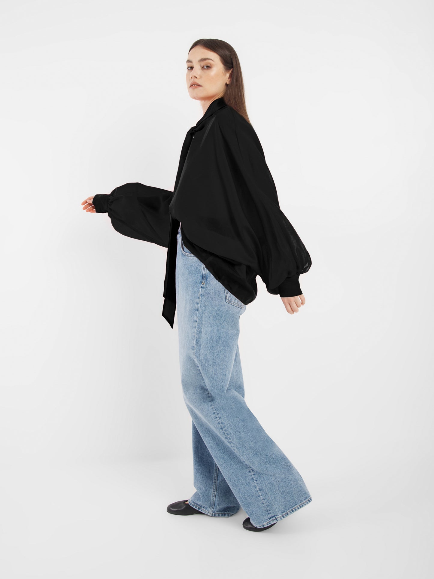 Eine Frau steht seitlich vor einem weißen Hintergrund. Sie trägt schwarze Antonia Bow Schuhe, eine lockere SOSUE Bluse mit voluminösen Ärmeln und hellblaue, weit geschnittene Jeans. Sie schaut mit neutralem Blick in die Kamera.