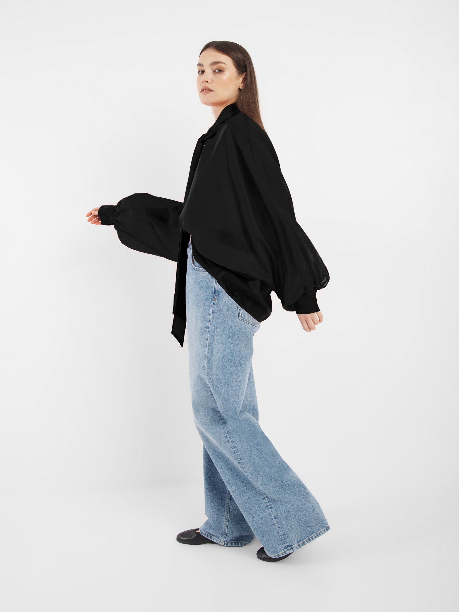 Eine Frau steht seitlich vor einem weißen Hintergrund. Sie trägt schwarze Antonia Bow Schuhe, eine lockere SOSUE Bluse mit voluminösen Ärmeln und hellblaue, weit geschnittene Jeans. Sie schaut mit neutralem Blick in die Kamera.