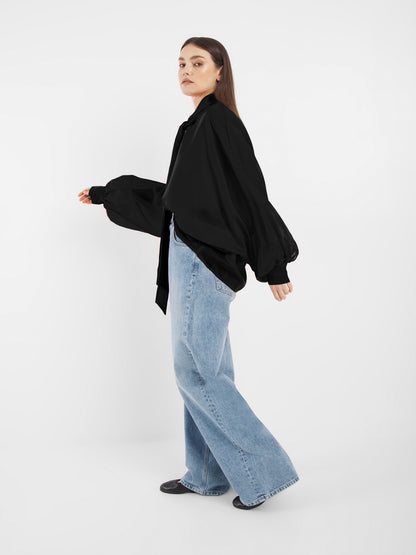 Eine Frau steht seitlich vor einem weißen Hintergrund. Sie trägt schwarze Antonia Bow Schuhe, eine lockere SOSUE Bluse mit voluminösen Ärmeln und hellblaue, weit geschnittene Jeans. Sie schaut mit neutralem Blick in die Kamera.