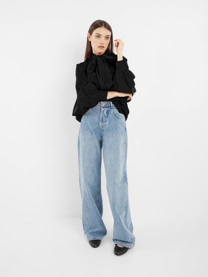 Eine Frau mit langen braunen Haaren steht vor einem weißen Hintergrund und trägt die schwarze Bluse Antonia Bow mit einer Schleife im Nacken, hellblaue Jeans mit weitem Bein und schwarze Schuhe. Sie blickt mit einer Hand neben ihrem Gesicht zur Seite.