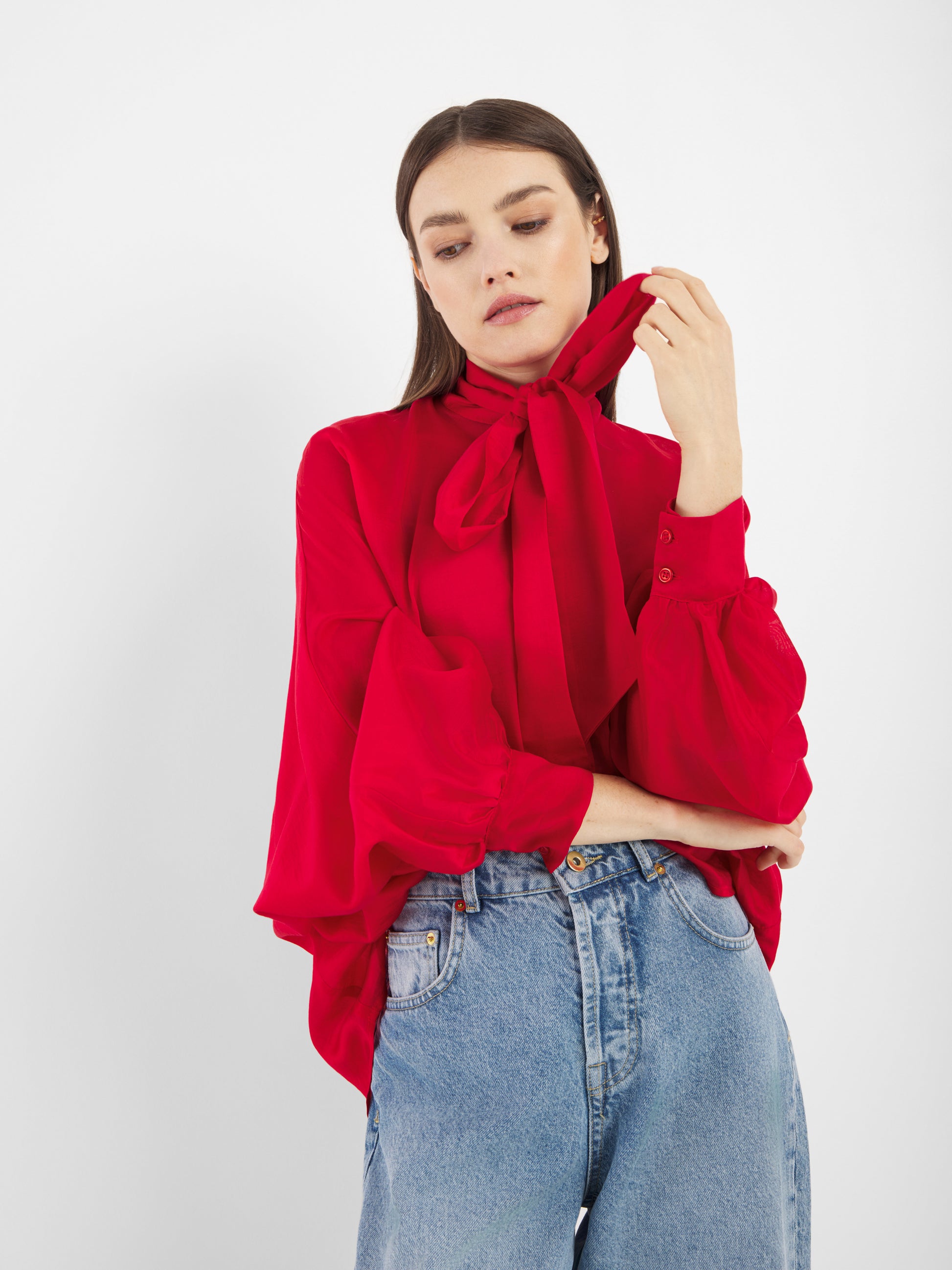Eine Frau trägt die Antonia Bow Red Damenbluse - eine leuchtend rote Lyocell-Bluse mit einer großen Schleife im Nacken und weiten Ärmeln - zusammen mit einer hoch sitzenden Bluejeans und posiert vor einem schlichten weißen Hintergrund.