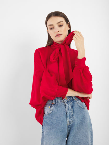 Die Bluse Antonia Bow Red mit ihrer auffälligen Schleife im Nacken und den weiten Ärmeln, kombiniert mit einer Bluejeans mit hoher Taille, wird von einer Person getragen, die vor einem weißen Hintergrund steht, nach unten schaut und die Schleife berührt.
