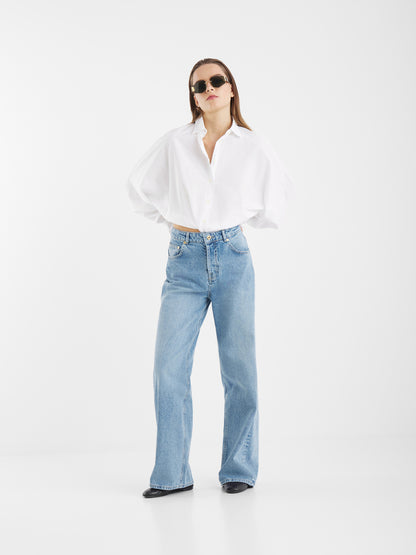 Eine Frau steht vor einem einfarbig weißen Hintergrund und trägt eine Sonnenbrille, die Bluse Antonia Slim White, die vorne umgeschlagen ist, eine hellblaue Jeans mit hoher Taille und weitem Bein sowie schwarze Schuhe. Die Arme hat sie hinter dem Rücken verschränkt.